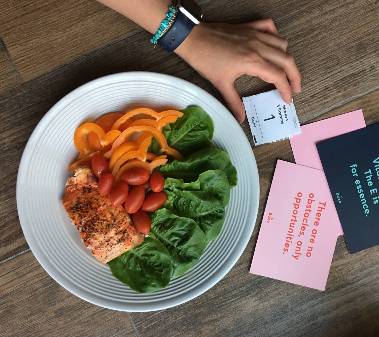 A female hand holding a daily supplement pack titled 'Maria's Vitamins' with a plate of colorful vegetables and salmon sitting in the background